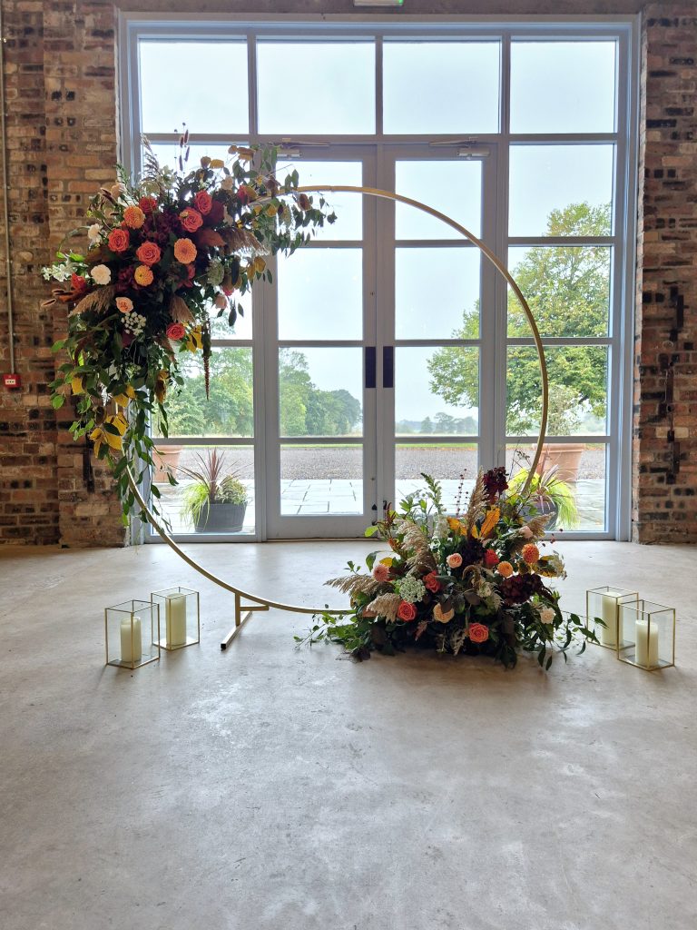 fraoch floral design large floral arrangement of pink and peach roses with greenery on a gold circular stand by a brick wall and glass doors; candles in glass lanterns on the floor nearby.