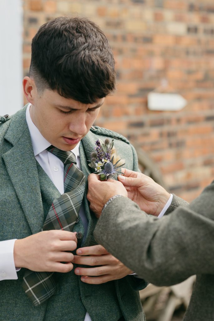 fraoch floral design young man in a green tweed suit watches as a boutonniere is pinned to his lapel by another person outdoors.